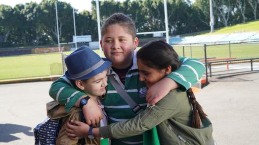 Von der Bank | Rechte: hr/Northern Pictures Production Jerry, Mikey und Salwa umarmen sich freundschaftlich fest im Rugbystadion vor dem Spielfeld. Alle drei tragen grüne Kleidung in der Mannschaftsfarbe des Bullfrog-Teams.