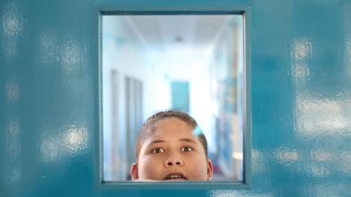 Süß und nicht fies | Rechte: hr/Northern Pictures Production Mikey, ein dunkelhaariger Junge mit zum Zopf gebundenen Haaren, schaut von aussen auf dem Flur durch das Sichtfenster einer verschlossenen Klassenzimmertüre ins Klassenzimmer hinein.