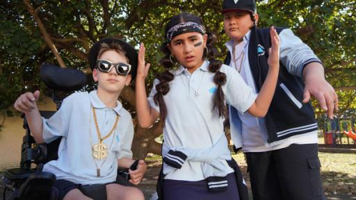 Das entscheidende Spiel | Rechte: hr/Northern Pictures Production Jerry, Salwa und Mikey haben sich für das Handballspiel auf dem Schulhof herausgeputzt. Jerry trägt eine fette, goldene Kette mit einem “J” und eine schwarze Sonnenbrille, Salwa hat sich Zöpfe geflochten und trägt ein blaues Bandana und Mikey hat ein Baseballcap auf und mehrere Goldketten um den Hals.