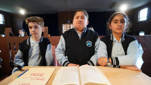 Kurzer Prozess | Rechte: hr/Northern Pictures Production Jerry, Mikey und Salwa sitzen in blauer Schuluniform auf einer Bank in einem düsteren Raum nebeneinander und schauen sehr ernst. Mikey hat seinen Mund nach unten gezogen. Vor ihnen auf dem Tisch liegt ein aufgeschlagenes Buch, im Hintergrund sitzen Mitschüler auf Sitzreihen.