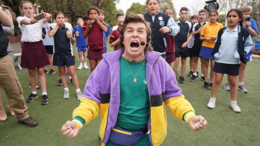 Der Workshop | Rechte: hr/Northern Pictures Production Der berühmte Handballspieler Gary Garrison kniet auf dem Rasen und wird umringt von Kindern, die ihn mit ihren Handies filmen. Er schreit aus vollem Hals, macht ein verzweifeltes Gesicht und breitet die Arme aus. Er hat ein grünes Sportshirt, eine bunte Fleecejacke an und ein Headset auf.