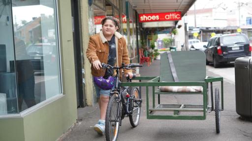 Mikey läuft in kurzer Jeanshose und brauner Cordjacke auf dem Gehweg an kleinen Geschäften vorbei und schiebt dabei ein Rad mit Seitenwagen, auf dem ein Sitz montiert ist. Er schaut entschlossen nach vorne.  