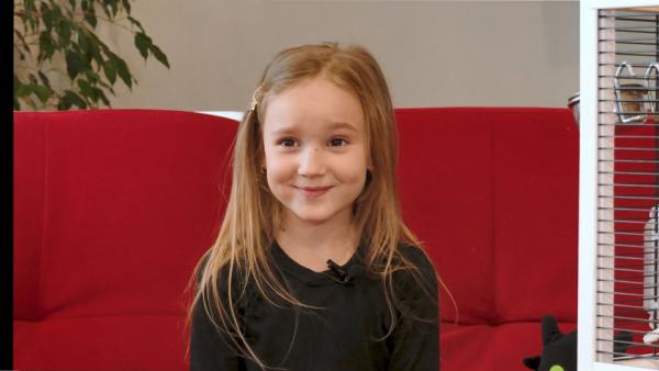 Elia sitzt auf einem roten Sofa. Sie hat lange blone Haare und lächelt.
