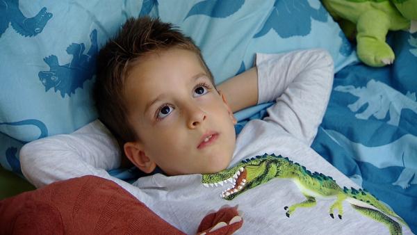 Marci liegt im Bett und hat seinen Arme hinter seinem Kopf verschränkt. Auf dem Kissen sind Dinos aufgedruckt.