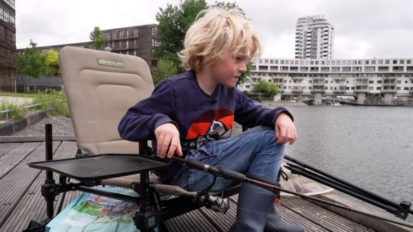 Louie sitzt an einem Bootssteeg auf einem Sessel. Mit der linken Hand hält er eine Angel ins Wasser.