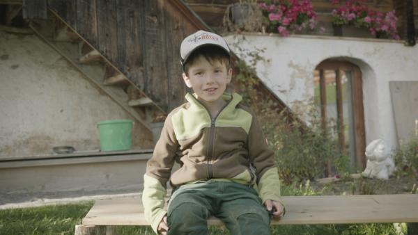 Fabio sitzt auf einer Holzbank vor einem Bauernhaus. Er trägt eine Mütze.