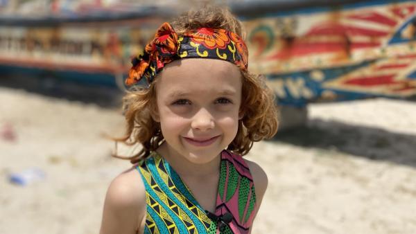 Ella steht am Strand vor einem bunten Boot. Sie trägt ein buntes Haarband und ein grünes Oberteil.
