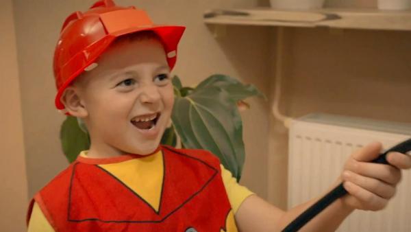 Roli im Feuerwehrkostüm mit Helm. Er hält einen Schlauch in der Hand und schaut begeistert.