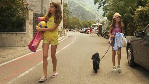 Rechte: ZDF/RTZG Marta geht mit Hund Beni an der Leine und ihre Schwester Anika auf einer Straße entlang. Sie tragen Badesachen. Rechts steht ein parkendes Auto.
