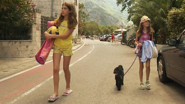 Marta geht mit Hund Beni an der Leine und ihre Schwester Anika auf einer Straße entlang. Sie tragen Badesachen. Rechts steht ein parkendes Auto.