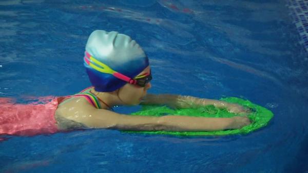 Liza schwimmt mit einem Schwimmbrett im Wasser. Sie trägt eine blaue Schwimmkappe.