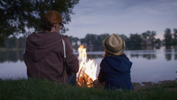 Bruno sitzt mit seinem Vater am Lagerfeuer mit Blick auf einen See. Es dämmert bereits.