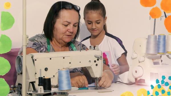 Das Mädchen Maša sitzt neben ihrer Oma, die mit einer Nähmaschine näht. Maša schaut ihr genau zu, denn sie möchte auch Nähen lernen.