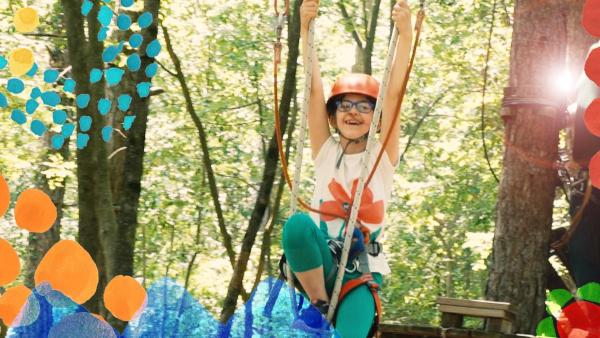 Nina mit Gurt und rotem Helm gesichert, hält sich hoch im Baum eines Kletterparks an Seilen einer Hängebrücke fest. Mit Freude im Gesicht hangelt sie sich zur gegenüberliegenden Seite.