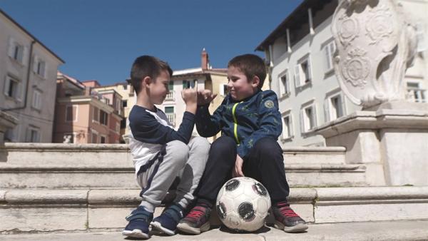 Die Freunde Elmin und Erik sitzen zusammen auf einer Treppe in der Stadt. Sie haben einen Fußball dabei, denn sie lieben Fußball.