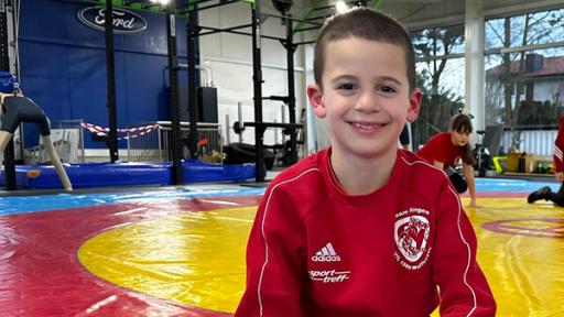 Erik ist Ringer | Rechte: hr Der 8-jährige Erik sitzt in einer Turnhalle. Im Hintergrund sind Turngeräte ufgestellt und rot-gelbe Turnmatten ausgelegt. Erik hat ganz kurze braune Haare trägt ein rotes Sweat-Shirt. Er lächelt in die Kamera.