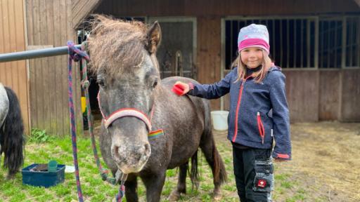 Nena und der Ausritt | Rechte: hr Nena steht neben ihrem Pony Molly. Sie hat eine Pferdebürste in der Hand und streicht über den Pferderücken. Im Hintergrund ist ein Pferdestall.