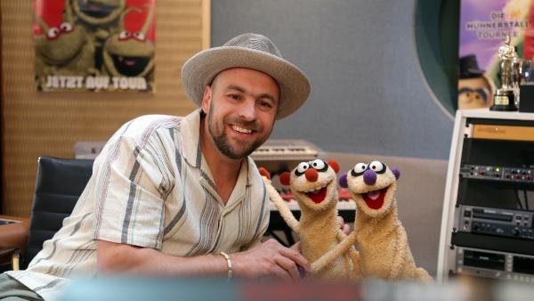 Max Mutzke sitzt auf einem Sessel in einem Musikstudio. Er neigt sich zu den Erdmännchen Jan und Henry und gibt Jan die Hand. Alle lächeln.