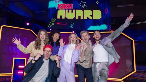 Gruppenfoto mit den Moderatorinnen und Moderatoren. Sie posieren und jubeln auf der großen Showbühne. Im Hintergrund prangt der Schriftzug "KiKA Award".