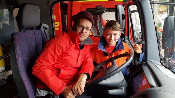 Ben sitzt in einer roten Jacke in einem Feuerwehrauto am Steuer. Paul schaut durch die offene Fahrertür in Auto.