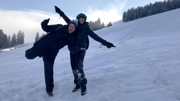 Auf einem Schnee bedeckten Hang stehen Ben und Jess und grinsen in die Kamera.