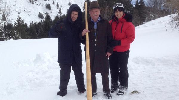 Im Schnee steht Ben links und Jess rechts. Zwischen ihnen steht ein Mann mit einem Alphorn. Im Hintergrund mit Schnee bedeckte Berge.