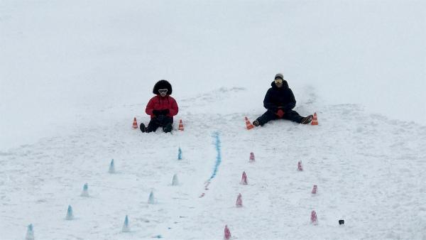 Im Schnee sitzen Jess und Ben mit Po-Rutschern. Vor ihnen ist ein Weg mit Pilonen markiert.