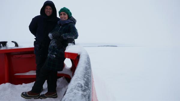 Ben und Jess stehen an der Reling auf einem Boot. Hinter ihnen ist ein zugefrorener See.