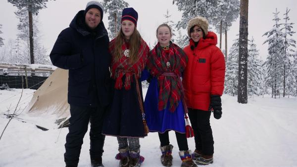 Im Schnee stehen von links nach rechts Ben, die Sami Mädchen Nadja und Sara und Jess ganz rechts. Nadja und Sara tragen traditionelle Sami-Trachten.