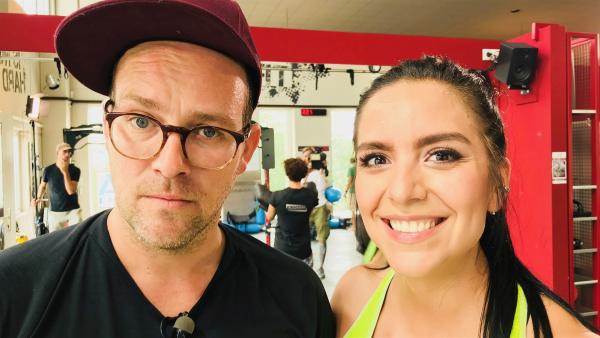 Groß die Köpfe von Ben, links, und Jess. Sie sind in einem Fitnesstudio. Im Hintergrund sieht man Sportgeräte und Leute, die daran trainieren.