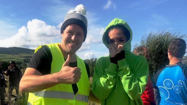 Ben und Jess werden frontal fotografiert. Jess hat einen Kapuzenpullover an und zieht die Kapuze fest zu. So verschwindet ihr Gesicht fast vollständig hinter dem Pullover. Ben hat eine grüne Warnweste an und hält den Daumen hoch.