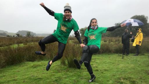 Ben und Jess sind auf der Wiese. Sie halten sich an der Hand und springen in die Luft. In diesem Moment wurde das Foto gemacht. Beide sind siegessicher in der Luft zu sehen. Man sieht, dass es geregnet hat. Ben und Jess sind etwas nass.