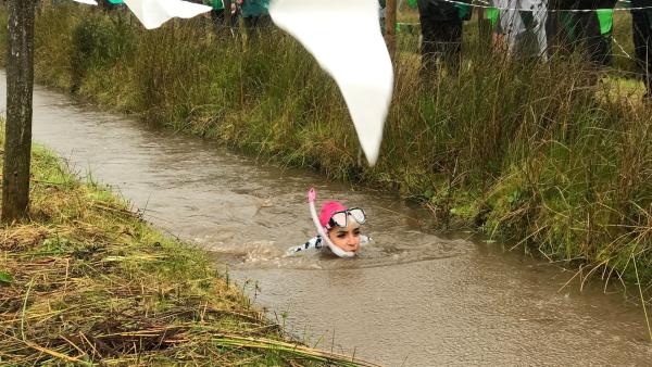 Jess gibt alles beim Sumpfschnorcheln durch 55 Meter Wassermatsch. Nur ihr Kopf und der Schnorchel gucken aus dem braunen Wasser. Im Hintergrund, am Rande des Sumpflaufes, sind Zuschauerinnen und Zuschauer mit Regenjacken zu sehen.