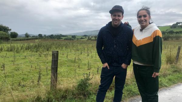 Ben und Jess stehen dicht beieinander, an einer umzäunten Wiese in Wales. Das Wetter ist nass und windig. Sie schauen beide in die Kamera und lächeln.