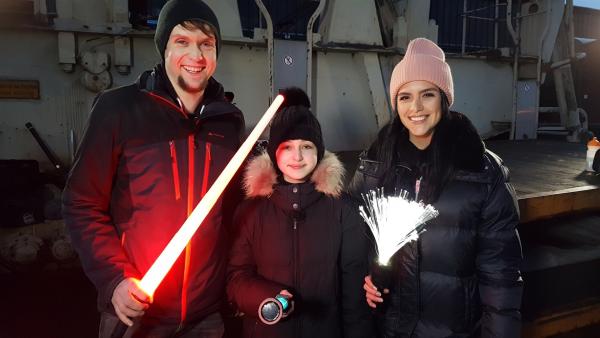 Jess steht rechts. Sie hält ein Bündel von leuchtenden Stäben in der Hand. Neben ihr steht ein Mädchen und ein Mann. Der Mann hat eine Art Laserschwert in der Hand.