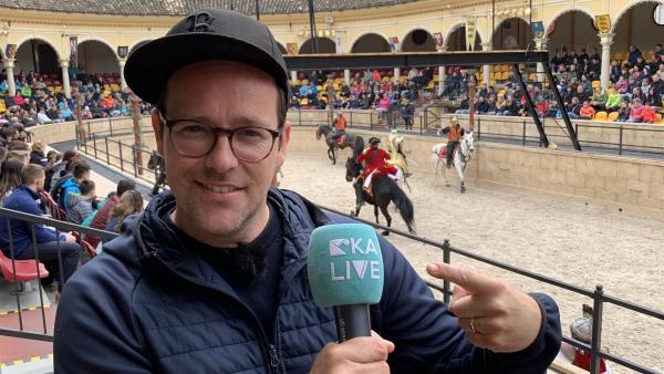 Ben mit Kappe, Brille und Mikrofon in der Hand, zeigt auf das Mikrofon mit der Aufschrift KiKA LIVE. Im Hintergrund eine historische Pferdeshow.