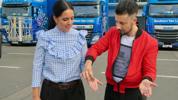 Auf einem Parkplatz stehen Jess und Andrew Basso. Jess hat die Hand geöfnnet, über welcher Andrew eine Münze hält.