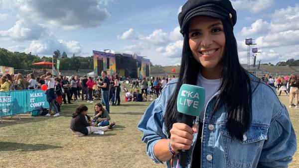 Vorne rechts steht Jess, die lächelt und ein 'KiKA LIVE' Mikrofon hält. Im Hintergrund ist das Gelände des Lollapalooza-Festival. Dort steht eine Bühne. Leute stehen und sitzen auf der Wiese.