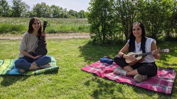 Jess und Leolixl sitzen nebeneinander auf einer Wiese und musizisieren mit ihren Ukulelen.