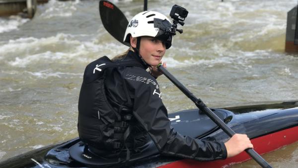 Christin, die Landesmeisterin im Wildwasserkajakfahren schaut vom Kajak aus in die Kamera. Sie hat ein Doppelpaddel in der Hand. Zur Sicherheit trägt sie einen Helm. An diesem ist eine Kamera befestigt. Außerdem hat sie einen Rucksack auf.