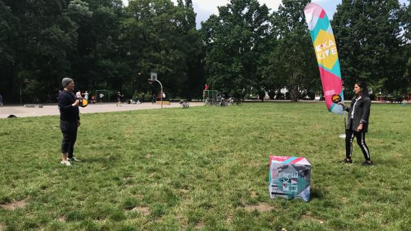 Ben und Jess testen auf der Wiese die besten Outdoorspiele. Sie sind halbweit fotografiert. Beide haben ein fristbee-ähnliches Spielgerät in der Hand. Im Hintergrund steht eine Fahne, im Vordergrund ein Würfel, beide mit dem Logo der Sendung KiKA-LIVE.