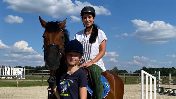 Es ist ein Reiterhof zu sehen. Jess sitzt auf einem Pferd. Emily hält das Pferd.