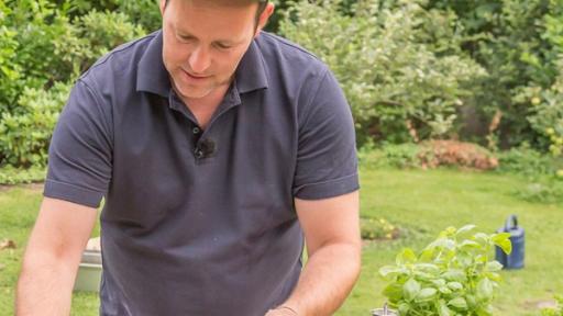 Ben und Ole bereiten das Essen für die Grillschale vor. Sie stehen beide am Tisch, auf dem die Lebensmittel und Küchenutensilien zu sehen sind. Im Hintergrund sieht man bereits das lodernde Feuer an einer Feuerstelle.