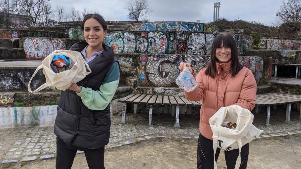 Links steht Jess, die ein Tüte mit Lebensmittel in die Kamera hält. Rechts steht Louisa Dellert, die auch eine Stofftüte mit Lebensmitteln hält. In ihrer rechent Hand hat sie ein Stück Plastik. Im Hintergrund eine mit Graffiti besprühte Steintreppe.