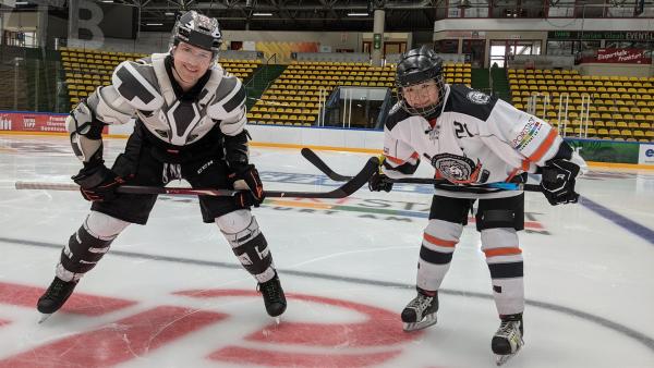 Auf der Eisfläche eines Eishockey-Stadions stehen Ben (links) und Lola. Sie tragen Eishockey-Ausrüstung, einen Helm und halten Hockey-Schläger in den Händen.