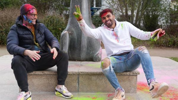 Ben und TikToker Younes Zarou sitzen in einem Park an einer Figur aus Stein. Beide sind mit bunten Pulver im Gesicht und auf der Kleidung bestreut und lachen.