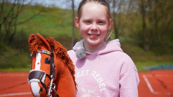 Die 12-jährige Emily ist ein Riesenfan von Hobby Horsing. Sie wird frontal fotografiert, trägt einen rosa Kapuzenpullover und hält das Steckenpferd in der Hand.
