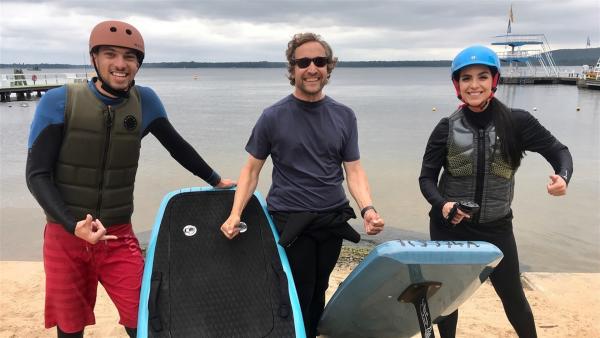 Jess, Finn und Bernd stehen im Sand des Berliner Müggelsees. Jess und Finn mit Helm und Wasseranzug. Bernd trägt eins der beiden Boards und hat eine schwarze Sonnenbrille auf.