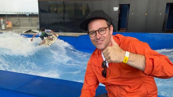 Vorne rechts sitzt Ben, der einen Daumen hoch in die Kamera zeigt. Hinter ihm ist ein Rapid-Surfing-Becken, in welchem Antonin auf einem Surfbrett eine Welle surft.