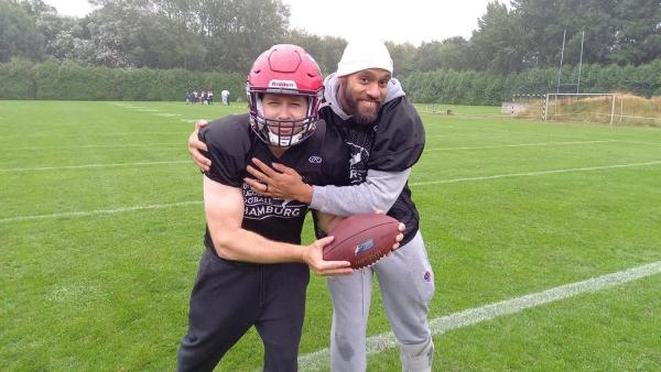 Am Rand eines Footballfelds steht Ben neben Footballer Kasim Edebali. Ben trägt Football-Montur, einen Helm und hält einen Football in der Hand.